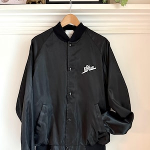 Puede incluir: Chaqueta de satén negra con cuello, puños y dobladillo acanalados negros. La chaqueta tiene una parte delantera con botones y dos bolsillos delanteros. El logotipo de Dr Pepper está impreso en blanco en la parte delantera. La chaqueta está colgada en una percha de madera.