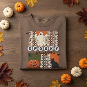 May include: A brown crewneck sweatshirt featuring a graphic print with a white ghost, a jack-o'-lantern, a black cat, and the text "SPOOKY SEASON" in white circular beads and brown script, surrounded by checkered and patterned vertical panels.