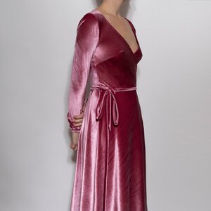 May include: A long, flowing wrap dress in a dusty rose velvet fabric. The dress features long sleeves, a V-neckline, and a waist tie. The skirt drapes to the floor, creating an elegant silhouette. The dress is a solid color, with a subtle sheen.