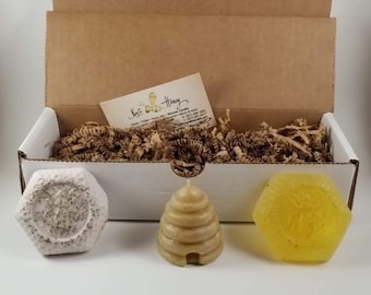 May include: A gift box containing three hexagonal candles and a small card. The candles are shaped like a honeycomb, a beehive, and a lavender-infused candle. The box is filled with brown crinkle paper. The card reads "Soft Gold Honey".