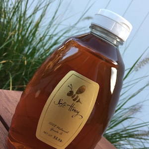 May include: A clear plastic bottle filled with amber-colored honey. The bottle has a white cap and a gold label with the words "Soft Honey" and a bee illustration. The label also states "100% Natural Grade A" and "NET WT. 32 oz."