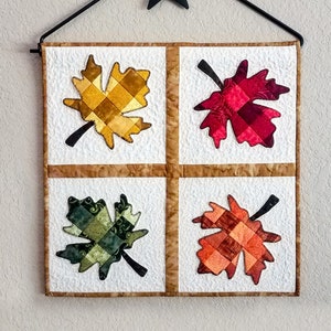 May include: A quilted wall hanging with four squares, each featuring a different colored maple leaf. The leaves are made of patchwork fabric in shades of red, orange, yellow, and green. The wall hanging is hung on a black metal star-shaped hook.