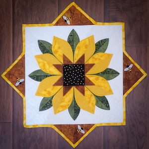 Op de afbeelding: Een witte gestikte tafelrunner met een geel zonnebloemontwerp en bruine bijen in de hoeken. De zonnebloem heeft een bruin centrum met witte stippen en gele bloemblaadjes.