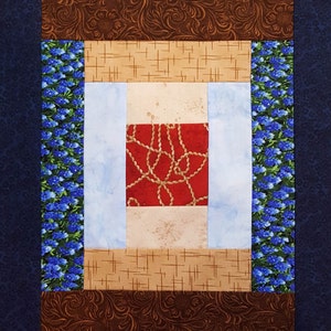 May include: A square quilt block with a central red square featuring a gold rope design. It is surrounded by light blue, tan, and brown patterned fabric. The edges have a blue floral pattern and a dark blue border.