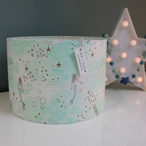 May include: A light green lampshade with a mermaid and star pattern. The lampshade has a white background with gold stars and dots. The text "Jack + Rose" is on a white tag attached to the lampshade.