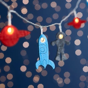 May include: A string of fairy lights with felt rocket, astronaut, and spaceship shapes. The lights are white and the shapes are blue, gray, and red.