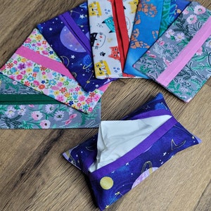 May include: A set of nine fabric tissue holders featuring a variety of patterns, including floral, cat, and galaxy designs. The holders have a purple trim and are arranged in a fan shape on a wooden surface. One holder is open, revealing a white tissue.