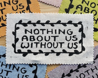 Può includere: Un cerotto di tessuto bianco con testo nero che recita "Nothing About Us Without Us" circondato da un bordo nero a foglie. Il cerotto è circondato da altri cerotti di tessuto con testo e disegni simili.