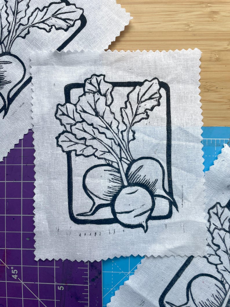 Puede incluir: Parches cuadrados de tela blanca con una impresi&oacute;n de linograbado negro de r&aacute;banos y hojas. El dise&ntilde;o del r&aacute;bano est&aacute; enmarcado dentro de un rect&aacute;ngulo negro. La tela tiene un borde en bruto sin terminar.