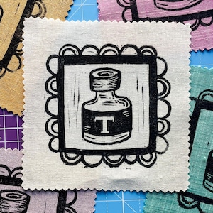 Peut inclure: Un patch de tissu carré blanc cassé avec une illustration d'encrier noir. La bouteille porte un "T" proéminent sur son étiquette et est encadrée d'une bordure festonnée. D'autres patchs de tissu avec des motifs similaires sont visibles en arrière-plan.