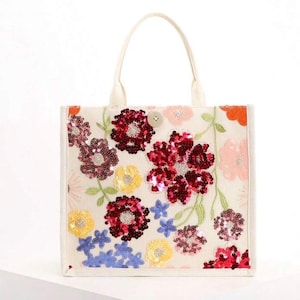 May include: A cream-colored tote bag featuring a vibrant floral pattern made of multi-colored sequins. The design includes red, yellow, pink, and blue flowers with green embroidered stems. The bag has two top handles and a single button closure.