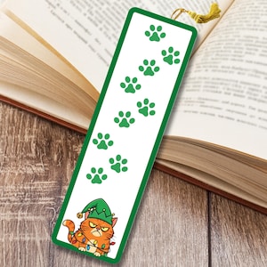 May include: A white bookmark with a green border and paw prints. The bottom of the bookmark features an orange cat wearing a green elf hat and Christmas lights. A gold tassel hangs from the top of the bookmark. The bookmark is placed in an open book.