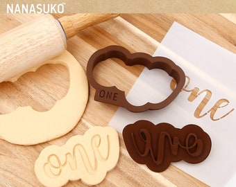 May include: A set of brown plastic cookie cutters in the shape of the word "one" on a wooden cutting board. The cutters are used to create cookies for a first birthday party.
