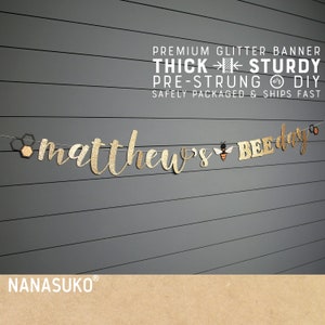 May include: Gold glitter banner with the text "matthew's BEE day" and a small bee illustration. The banner is pre-strung and comes with a  "THICK STURDY" and "PRE-STRUNG or DIY" description. The banner is safely packaged and ships fast.