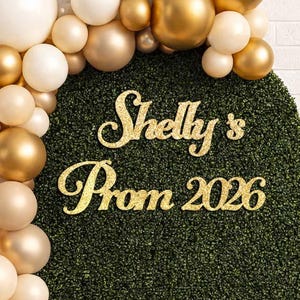 May include: A decorative prom backdrop featuring a green hedge-like surface with gold glitter lettering that reads "Shelly's Prom 2026." A balloon garland in shades of white, gold, and beige arches over the top and left side of the backdrop.
