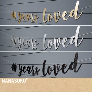 May include: Three banners with the text "40 years loved" in gold, silver, and black glitter. The banners are strung on white twine and are hanging against a grey background.
