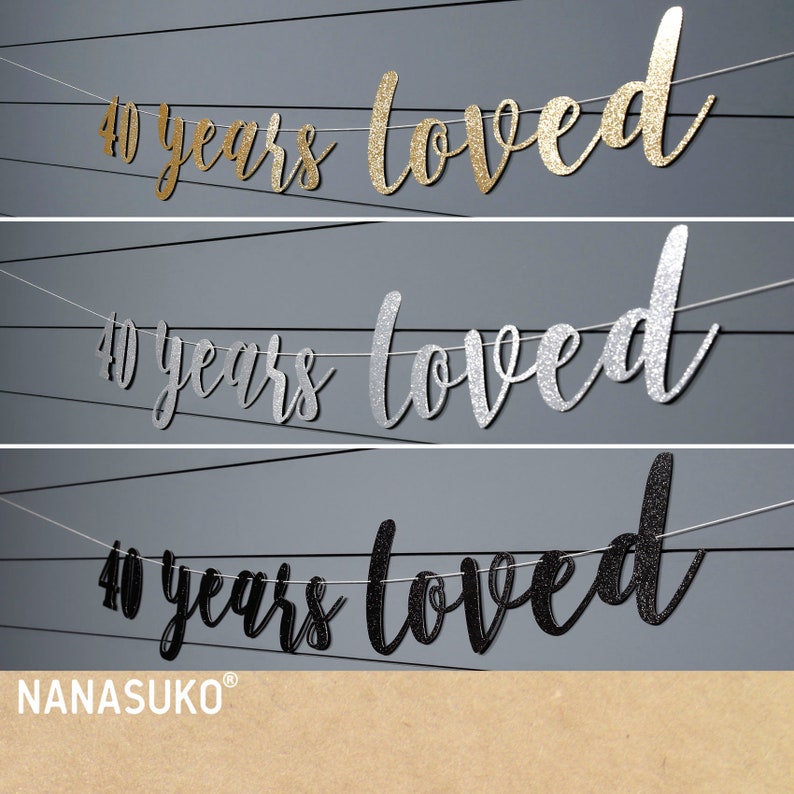 May include: Three banners with the text "40 years loved" in gold, silver, and black glitter. The banners are strung on white twine and are hanging against a grey background.