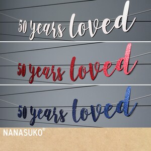 May include: Three banner decorations with the text "50 years loved" in white, red, and blue glitter. The banners are strung on white string.