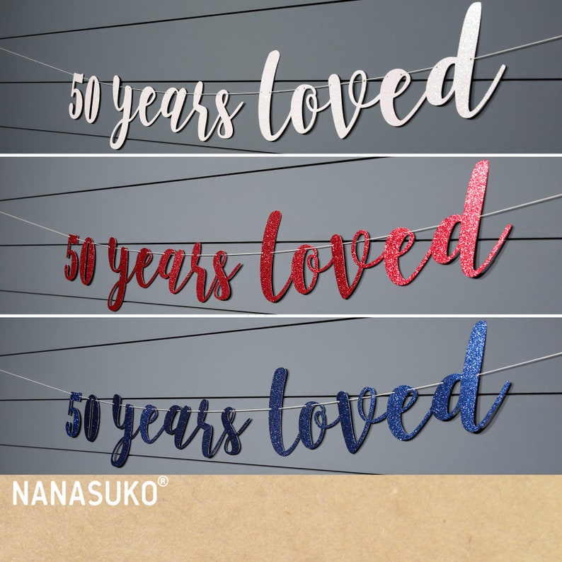 May include: Three banner decorations with the text "50 years loved" in white, red, and blue glitter. The banners are strung on white string.