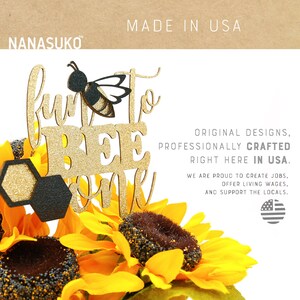 May include: Fun to Bee One cake topper with a glitter gold finish and a black and yellow bee. The topper is sitting on a bed of sunflowers.