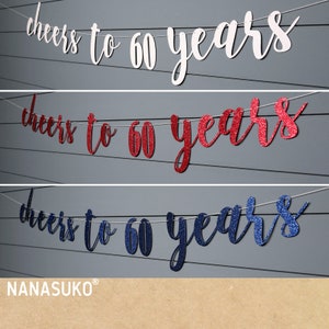 May include: Three banners with the text "cheers to 60 years" in a cursive font. The top banner is white, the middle is red glitter, and the bottom is blue glitter. The banners are strung on a thin white string.