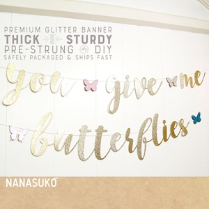 May include: Gold glitter banner with the text "you give me butterflies". The banner is strung with pink, blue, and gold glitter butterflies.