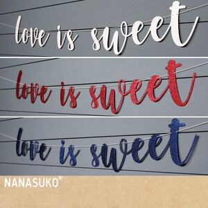 Puede incluir: Tres pancartas con el texto "love is sweet" en blanco, rojo con purpurina y azul con purpurina. Las pancartas est&aacute;n colgadas de una cuerda.