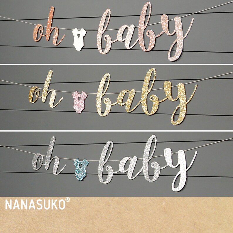 May include: Three banners with the text "oh baby" in gold, rose gold, and silver glitter. Each banner has a small white, pink, or blue onesie hanging from it.