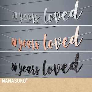 May include: Three 80 years loved banners in silver, rose gold, and black glitter. The banners are strung on white string and are perfect for an 80th birthday party.