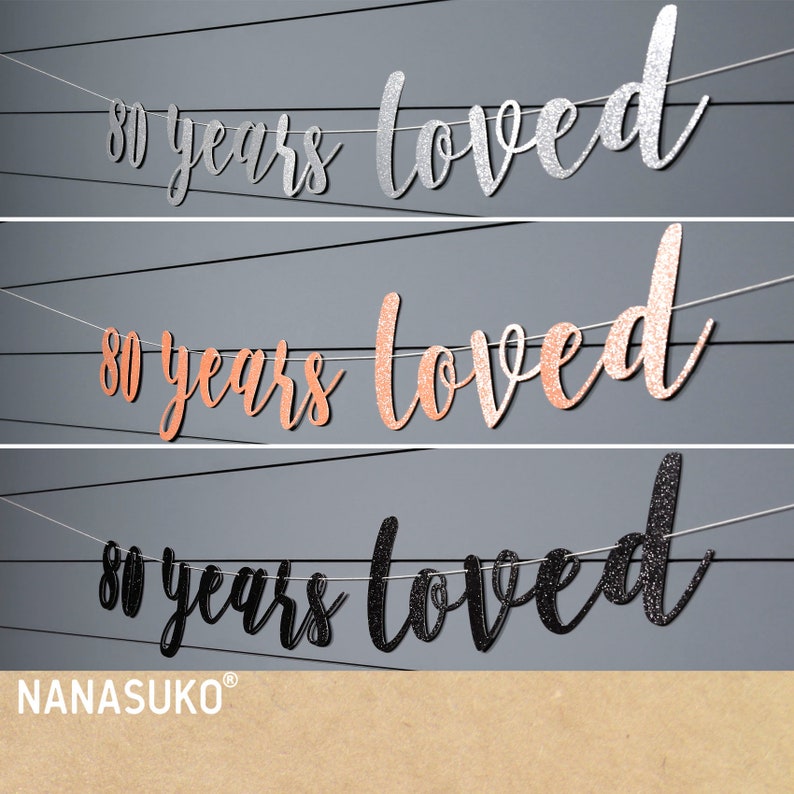 May include: Three 80 years loved banners in silver, rose gold, and black glitter. The banners are strung on white string and are perfect for an 80th birthday party.