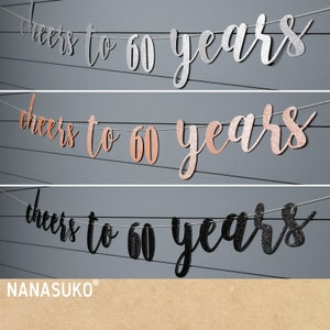 May include: Three banners with the text "cheers to 60 years" in silver, rose gold, and black glitter. The banners are strung on white string and are against a gray wooden background. The brand name "NANASUKO" is at the bottom.