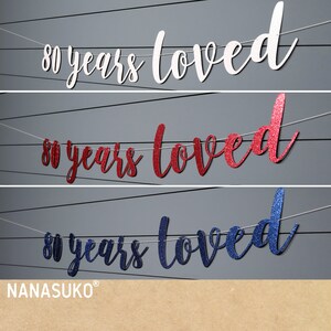 May include: Three banner decorations with the text "80 years loved" in white, red glitter, and blue glitter. The banners are strung on white string.