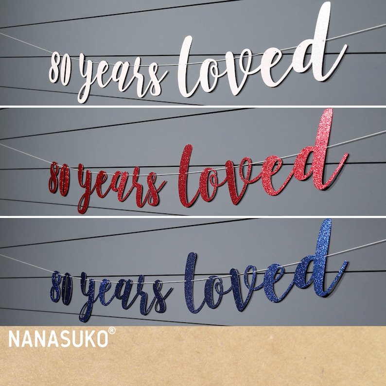 May include: Three banner decorations with the text "80 years loved" in white, red glitter, and blue glitter. The banners are strung on white string.