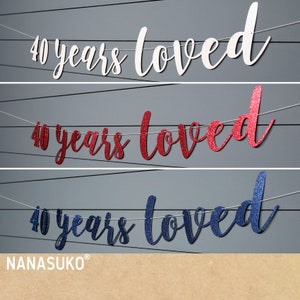 May include: Three banner decorations with the text "40 years loved" in white, red glitter, and blue glitter. The banners are strung on white twine and are hanging against a grey background.