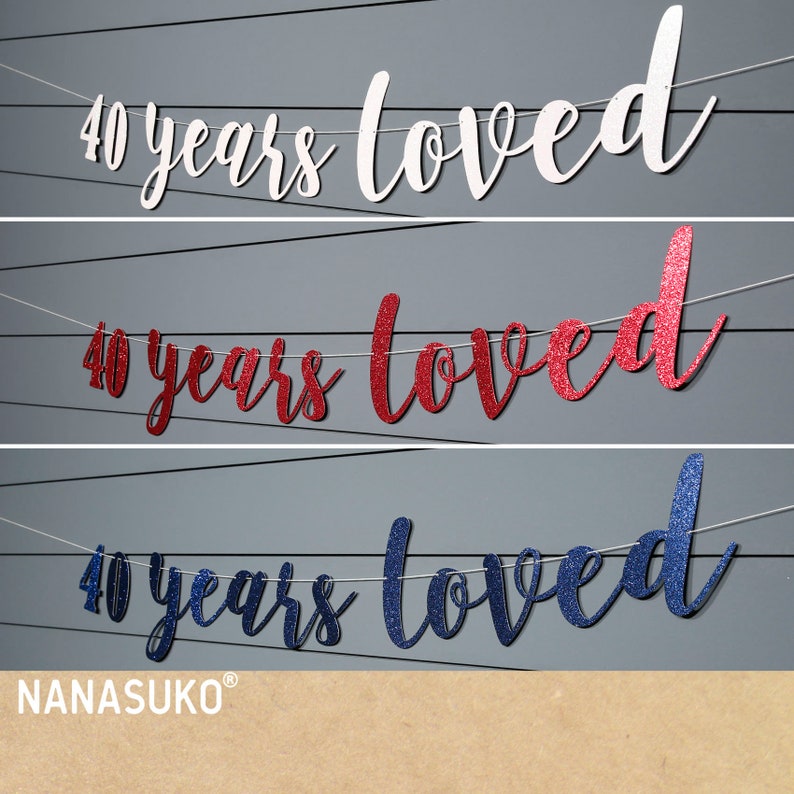 May include: Three banner decorations with the text "40 years loved" in white, red glitter, and blue glitter. The banners are strung on white twine and are hanging against a grey background.