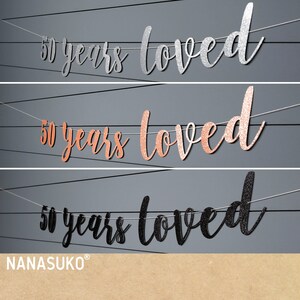 May include: Three banner decorations with the text "50 years loved" in silver, rose gold, and black glitter. The banners are strung on white twine.