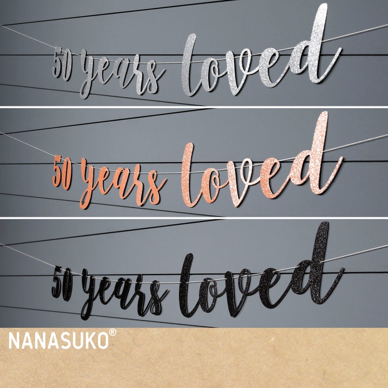 May include: Three banner decorations with the text "50 years loved" in silver, rose gold, and black glitter. The banners are strung on white twine.