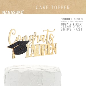 May include: Gold glitter cake topper with the text "Congrats Lauren" and a black graduation cap. The topper is on a white frosted cake.