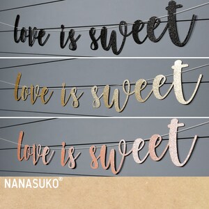 Puede incluir: Tres pancartas de "Love is Sweet" en purpurina negra, dorada y rosa dorada. Cada pancarta est&aacute; enhebrada con hilo blanco y tiene una fuente cursiva.