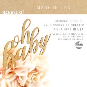 Puede incluir: Topper de pastel de madera dorada con las palabras "oh baby" en una fuente cursiva. El topper est&aacute; sentado encima de un ramo de flores de color rosa p&aacute;lido. El fondo es una superficie blanca con el texto "MADE IN USA" y un mensaje sobre el apoyo a las empresas locales.