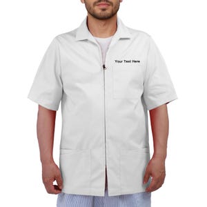 Pode incluir: Camisa branca de manga curta com zíper e dois bolsos frontais. A camisa tem gola e o texto "Your Text Here" está impresso no peito. A camisa é usada por uma pessoa.