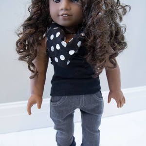 May include: A doll with brown curly hair wearing a black shirt with a white polka dot scarf, gray pants, and black shoes.