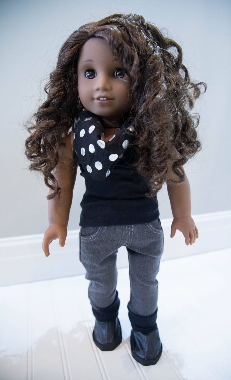 May include: A doll with brown curly hair wearing a black shirt with a white polka dot scarf, gray pants, and black shoes.