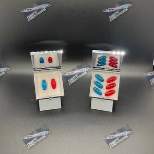 Peut inclure: Deux petites boîtes argentées ouvertes présentent chacune plusieurs faux ongles ovales et brillants. Une boîte contient des ongles bleus et rouges, l'autre contient des ongles bleus et rouges. Le fond est noir.