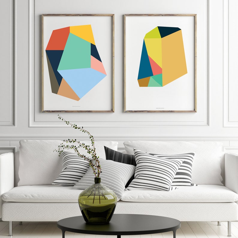May include: Two framed abstract geometric art prints with colorful shapes in shades of blue, green, yellow, orange, and red. The prints are hung on a white wall above a white couch with black and white striped pillows.