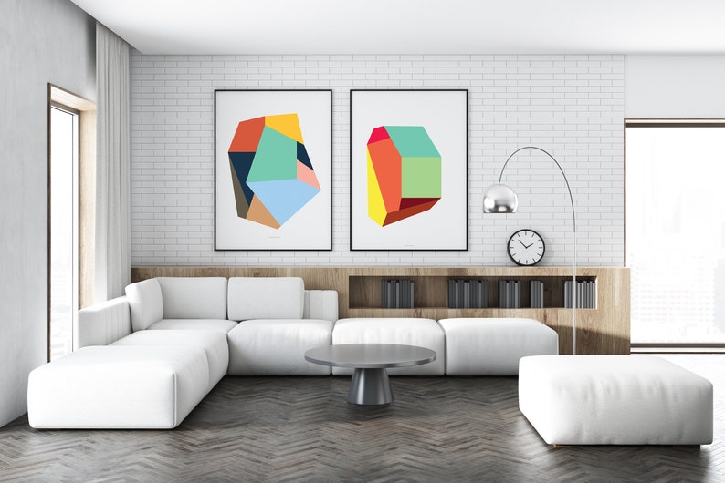 May include: Two abstract geometric art prints with colorful shapes in a modern living room with a white sectional sofa, a gray coffee table, and a white ottoman.