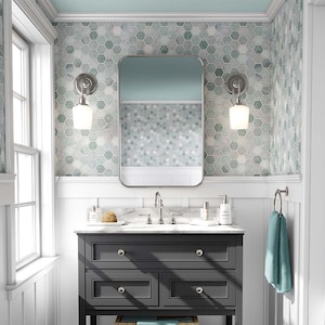 Puede incluir: Un tocador de baño de color gris oscuro con tres cajones y un estante inferior. El tocador tiene una encimera blanca con un lavabo y grifos cromados. El tocador está frente a una pared con zócalo blanco y un patrón de azulejos hexagonales de color azul claro y gris. Hay dos lámparas de pared con pantallas de vidrio blanco.