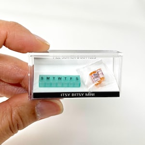 Pode incluir: Um organizador de pílulas em miniatura e duas garrafas de pílulas em miniatura em uma vitrine de acrílico transparente. O organizador de pílulas é turquesa e tem os dias da semana etiquetados: S M T W T F S. As garrafas de pílulas são laranja e têm etiquetas brancas. A vitrine tem uma base preta com o texto "ITSY BITSY MINI" impresso nela.