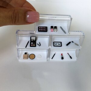 May include: A clear plastic makeup organizer with multiple shelves. The organizer contains miniature makeup items, including eyeshadow palettes, lipstick, nail polish, mascara, and eyeliner.