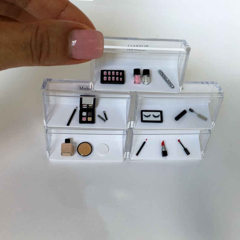 May include: A clear plastic makeup organizer with multiple shelves. The organizer contains miniature makeup items, including eyeshadow palettes, lipstick, nail polish, mascara, and eyeliner.